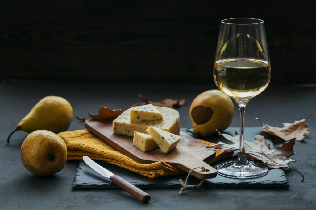 A glass of white wine served with cheese in a cutting board on dark background